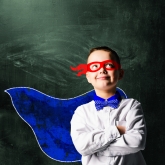 school boy wearing a superhero costume with blackboard behind him